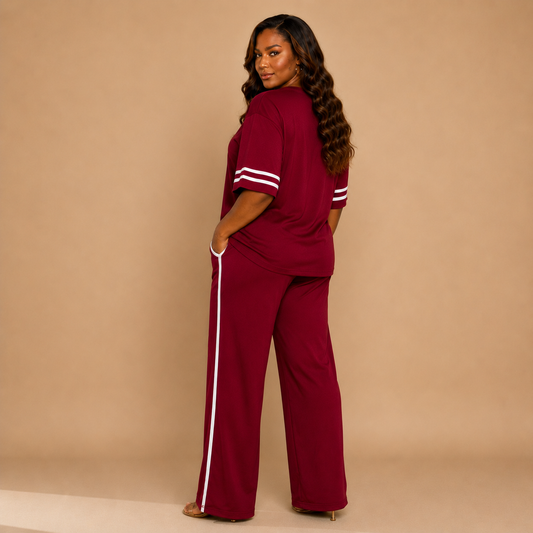 A woman modeling a matching burgundy two-piece lounge set with white stripe accents on the sleeves and down the side of the wide-leg pants, photographed against a solid beige studio backdrop.