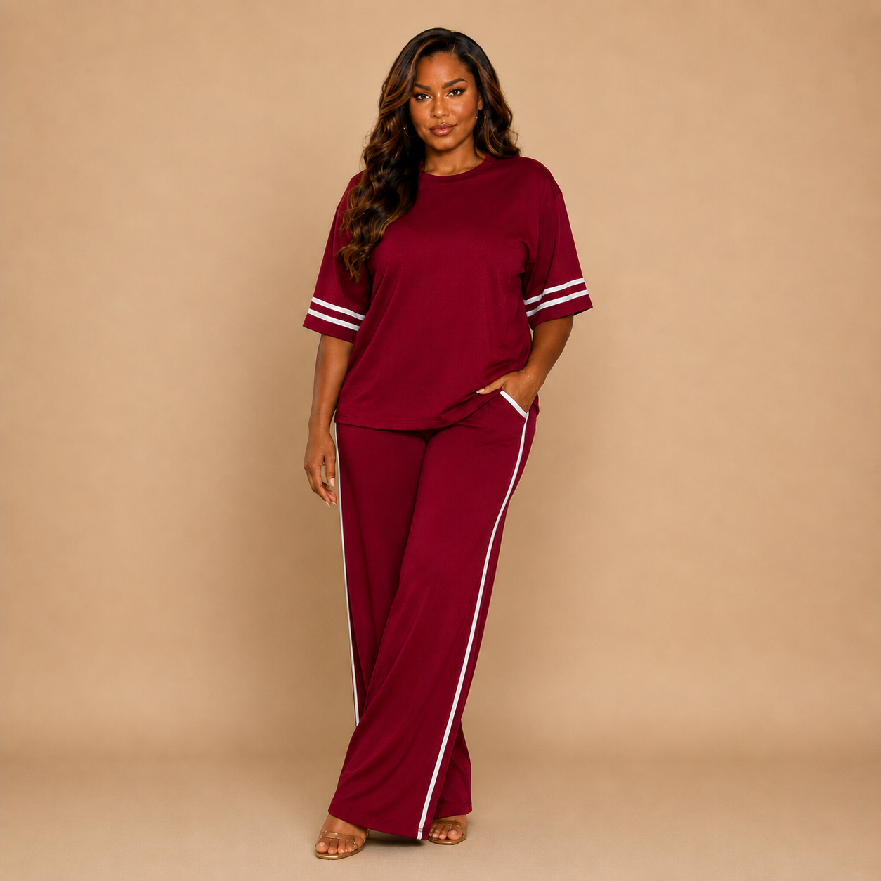 A woman modeling a matching two-piece lounge set in deep burgundy, featuring a short-sleeve top with white stripe details on the sleeves and matching wide-leg pants with white side stripes, set against a solid beige studio background.
