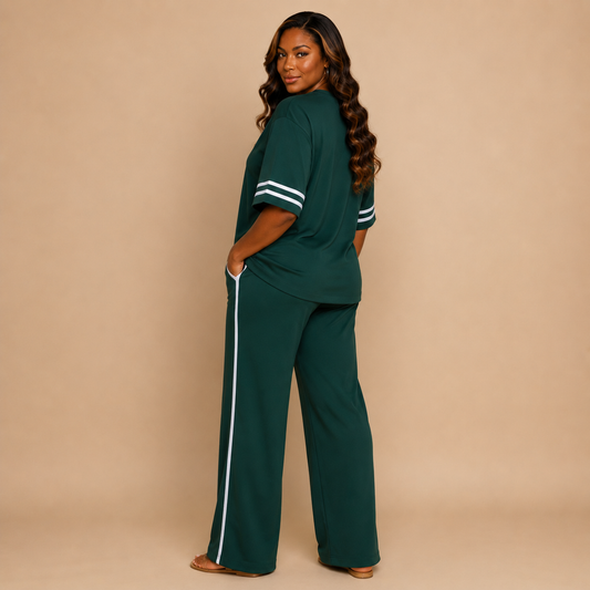 A woman modeling a dark green two-piece lounge set consisting of a short-sleeve top with white stripes on the sleeves and matching wide-leg pants with a white side stripe, set against a solid beige studio background.