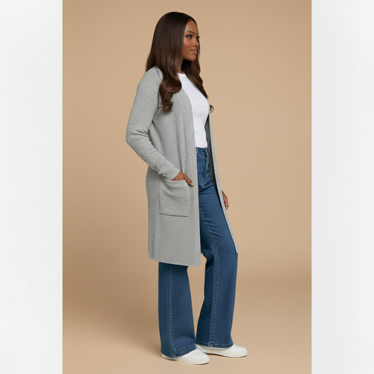 Woman wearing a long gray cardigan, white shirt, blue jeans, and white sneakers on a plain background