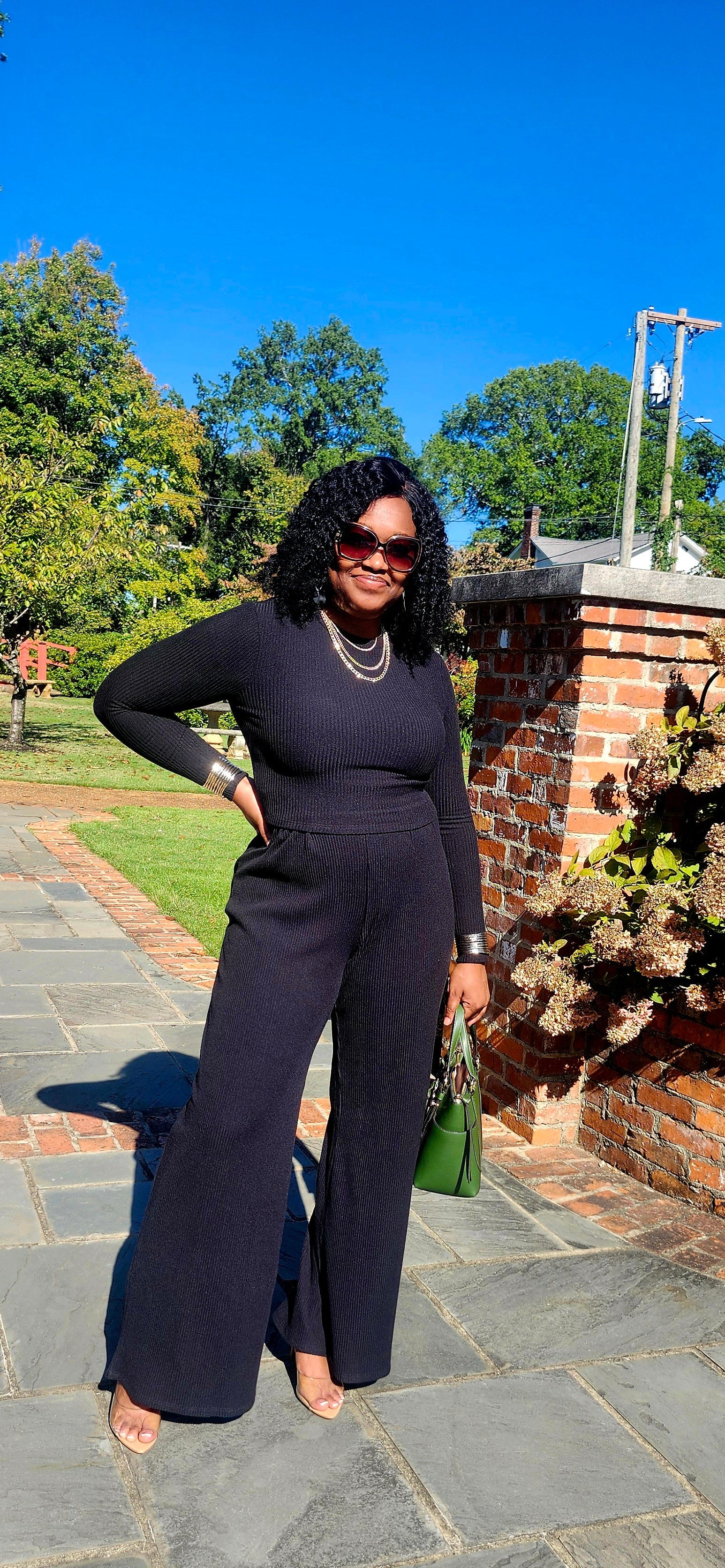 Woman in a black outfit standing on a stone path with trees and a brick wall in the background