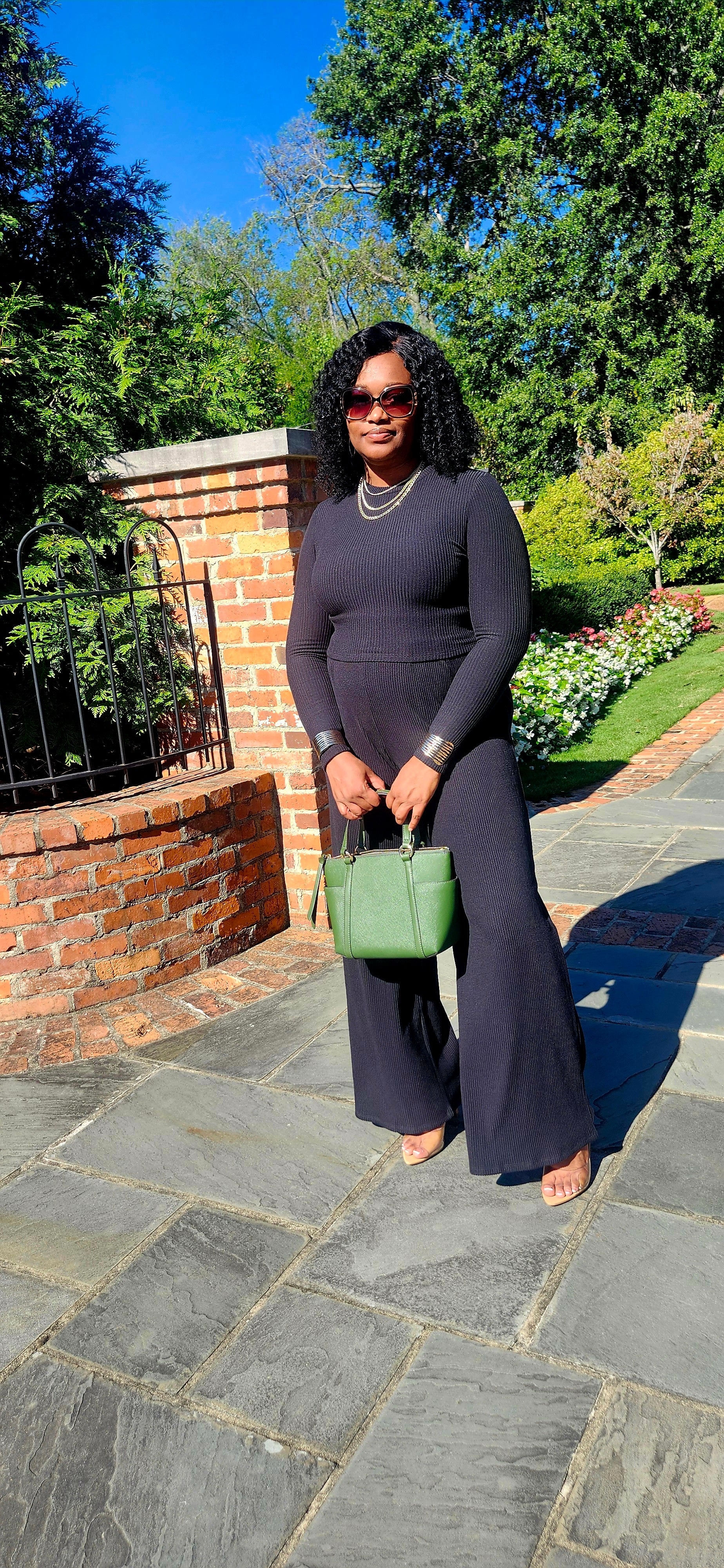 Woman in a black outfit holding a green handbag on a stone path with trees in the background