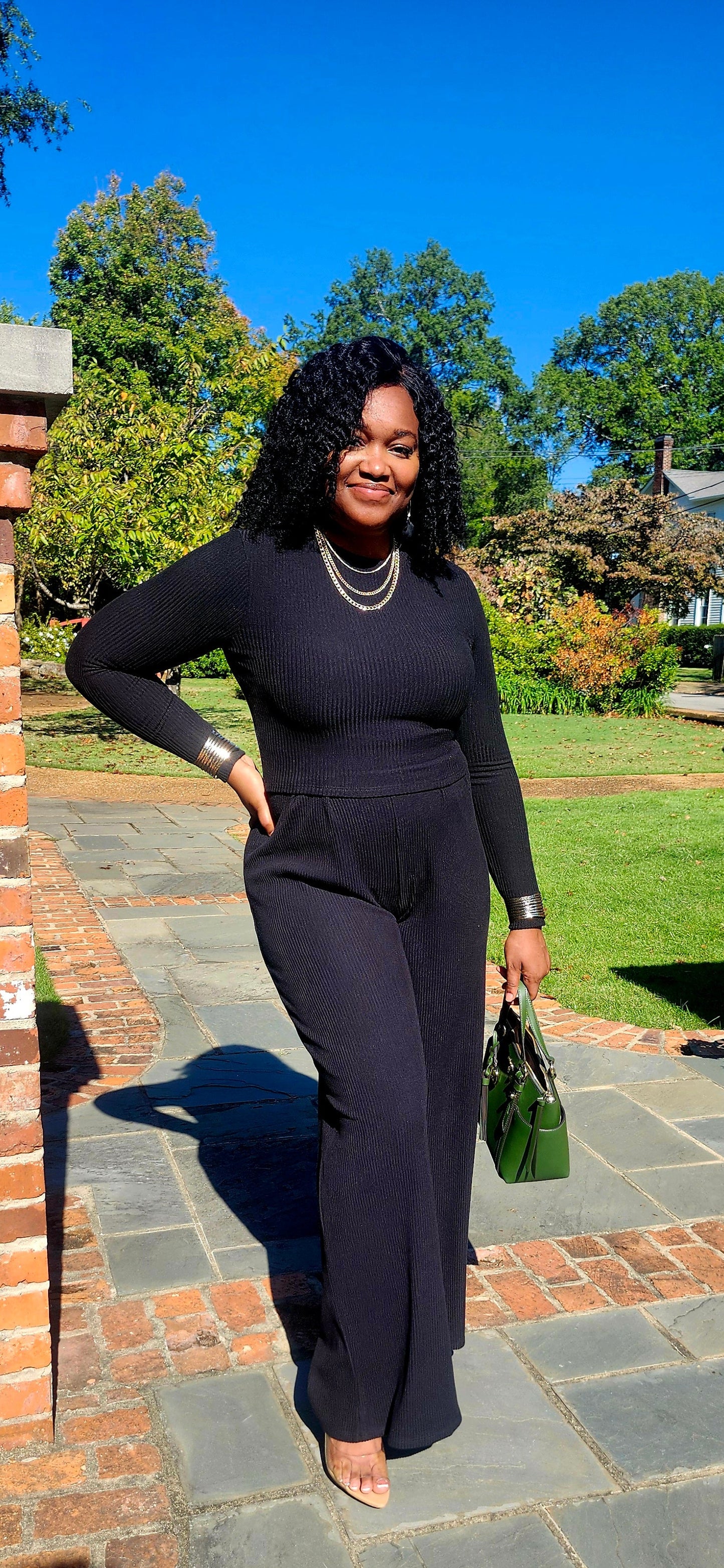Woman in a black outfit standing on a stone path with greenery and a clear blue sky in the background.