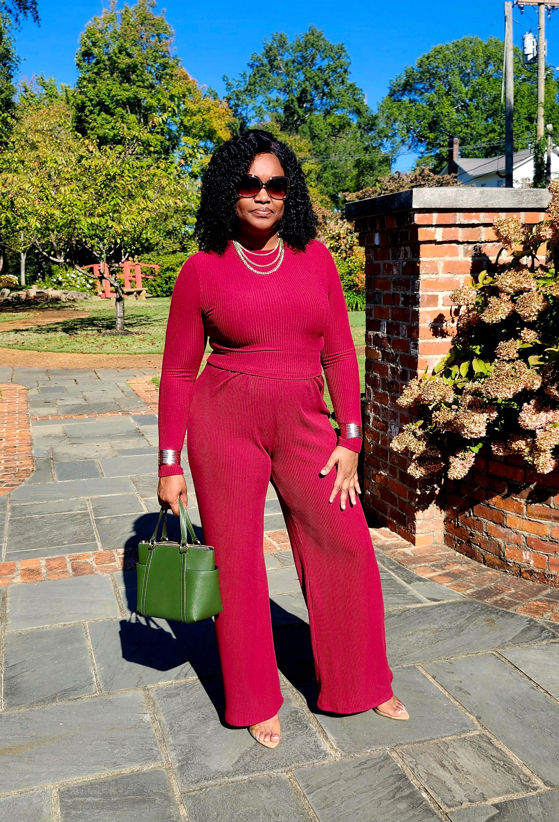 Woman in a red outfit standing outdoors on a stone path with trees and a building in the background.