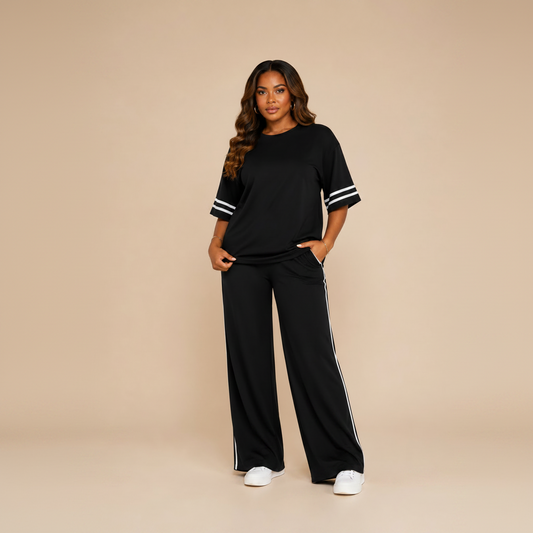 A woman modeling a black two-piece lounge set consisting of a short-sleeve top with white stripe accents on the sleeves and matching wide-leg pants with white side stripes, set against a solid beige studio background.