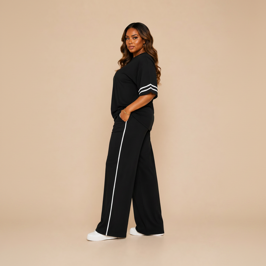 A woman modeling a black two-piece lounge set consisting of a short-sleeve top with white stripes on the sleeves and matching wide-leg pants with a white side stripe, set against a solid beige background.