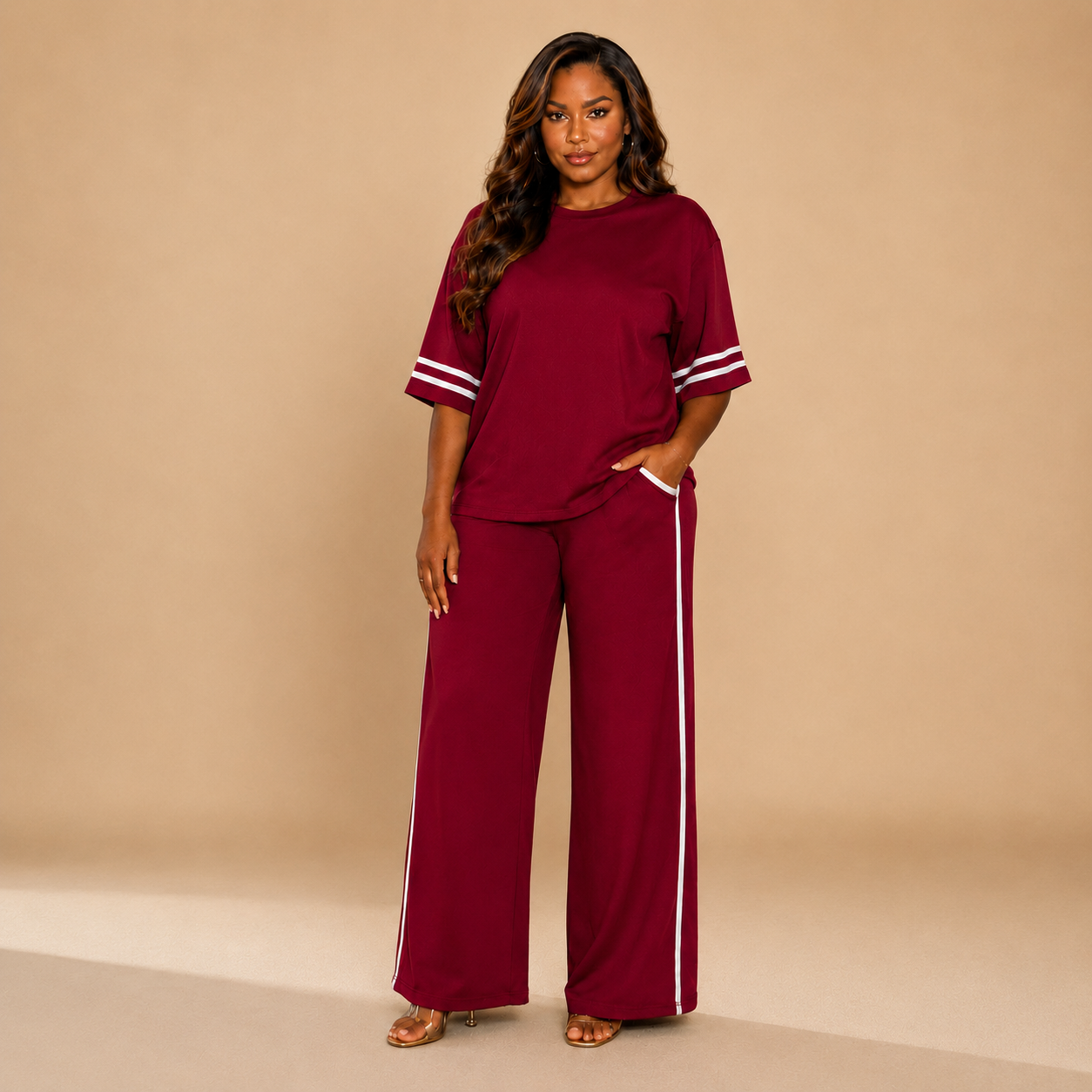 A woman modeling a matching burgundy lounge set consisting of a short-sleeve top with white stripe details on the sleeves and a pair of wide-leg pants with white side stripes, set against a solid beige studio background.