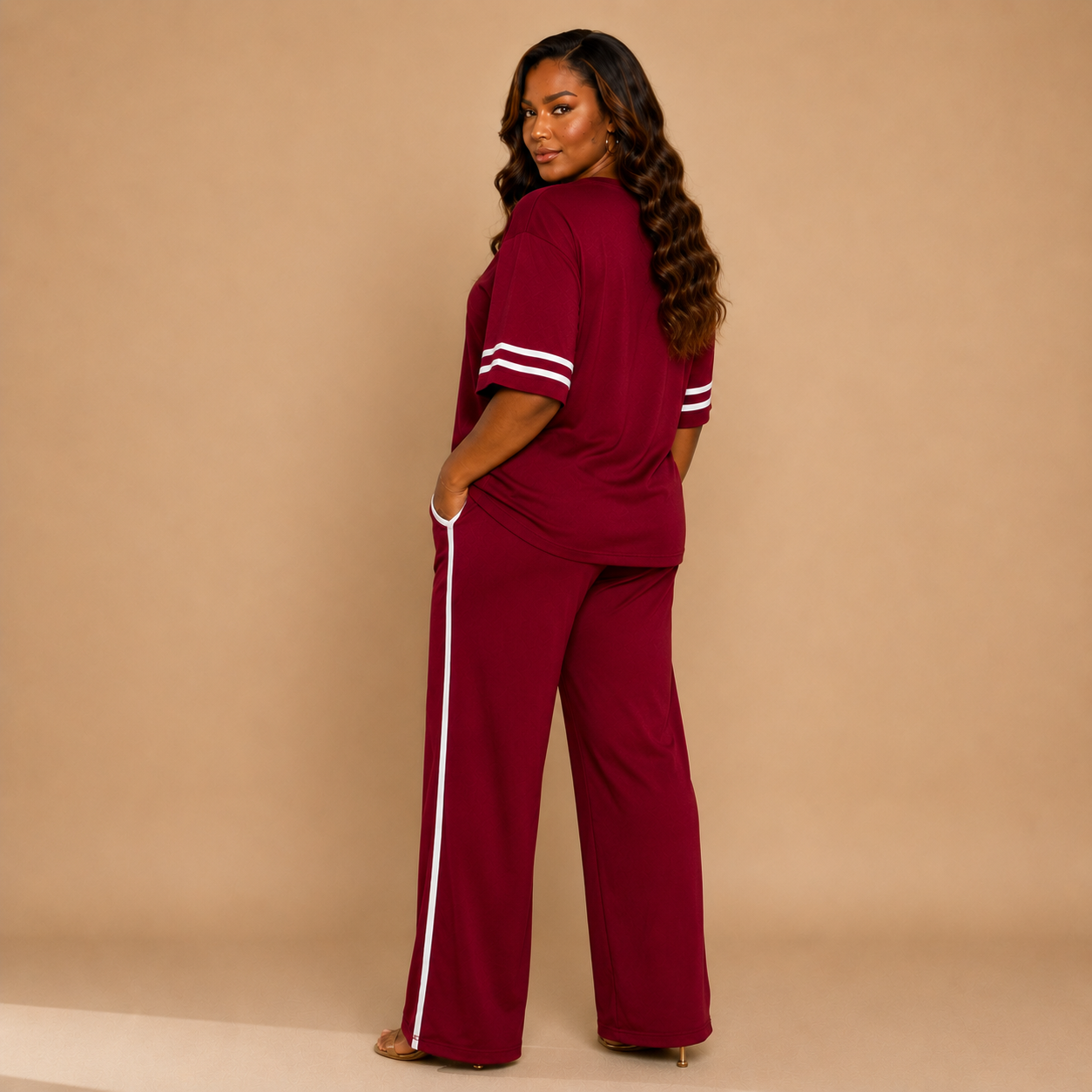 A woman modeling a matching burgundy two-piece lounge set with white stripe accents on the sleeves and down the side of the wide-leg pants, photographed against a solid beige studio backdrop.