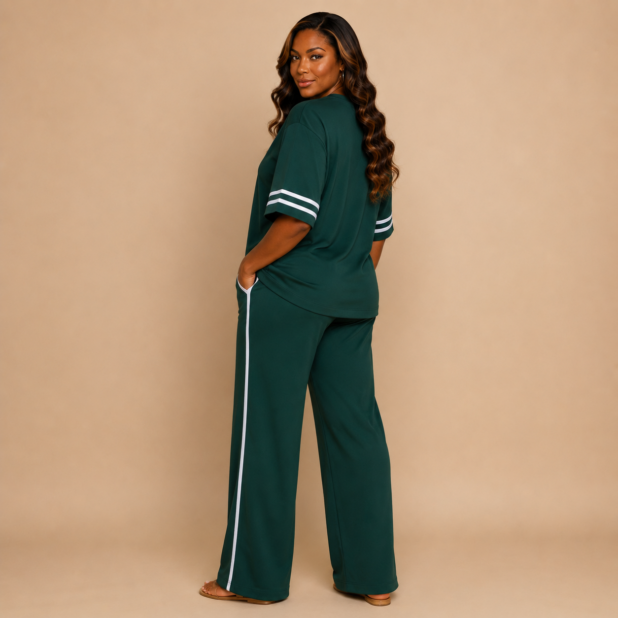 A woman modeling a dark green two-piece lounge set consisting of a short-sleeve top with white stripes on the sleeves and matching wide-leg pants with a white side stripe, set against a solid beige studio background.