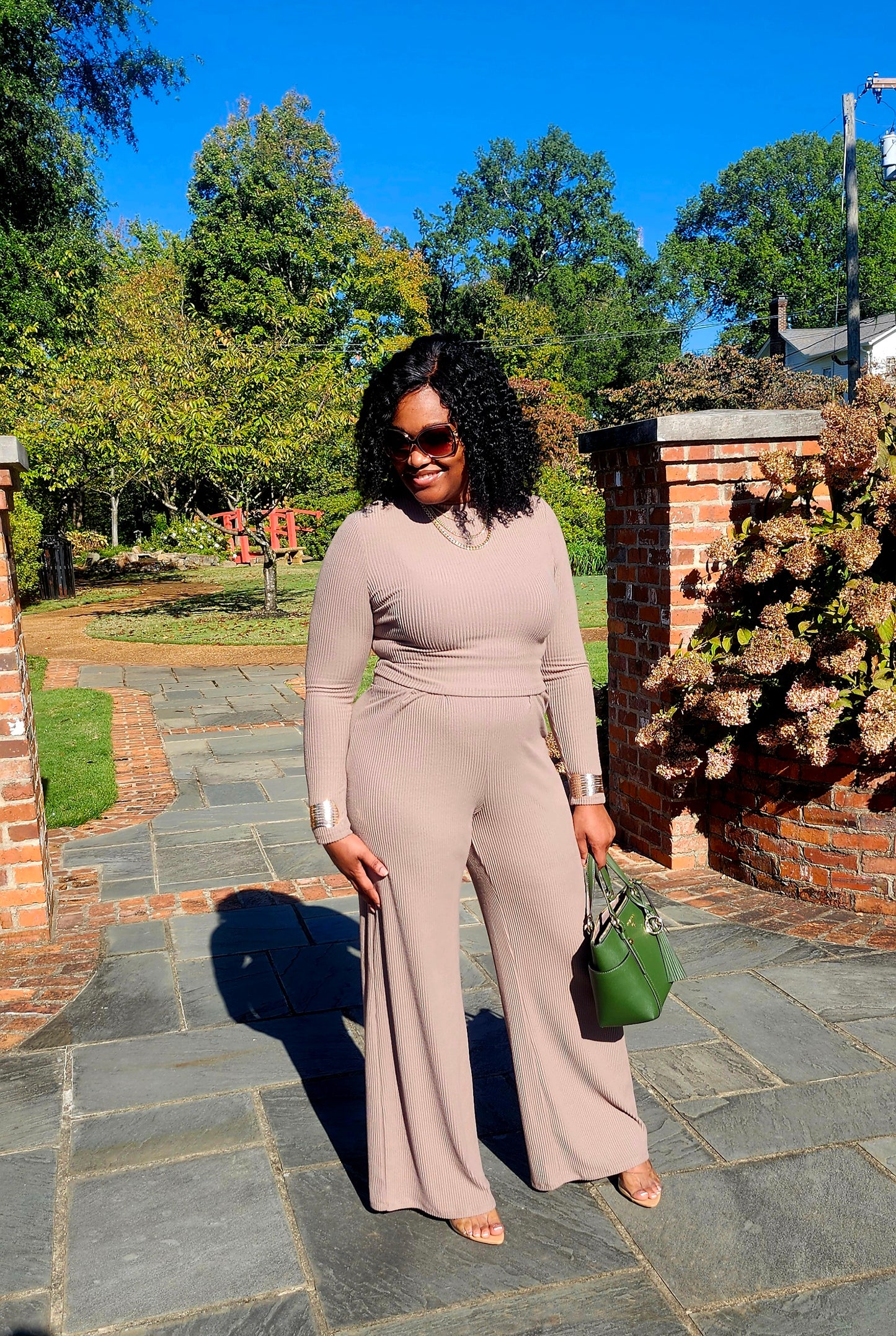 Woman in a beige jumpsuit standing outdoors on a stone path with trees and a clear blue sky in the background.