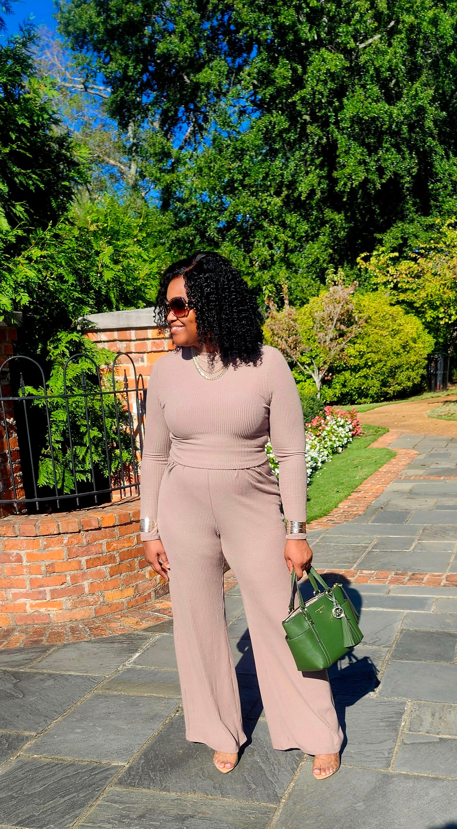 Woman in a beige jumpsuit holding a green handbag on a stone path with trees in the background
