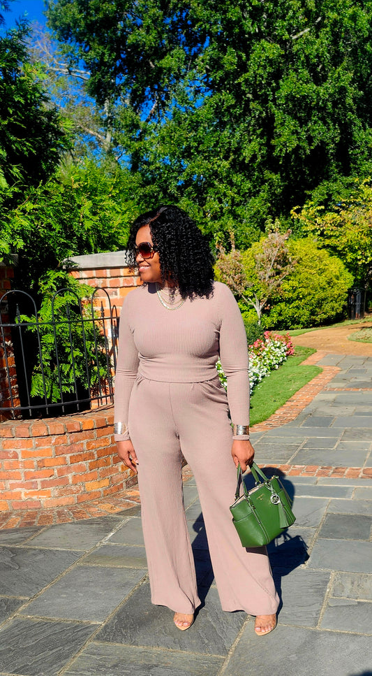 Woman in a beige jumpsuit holding a green handbag on a stone path with trees in the background