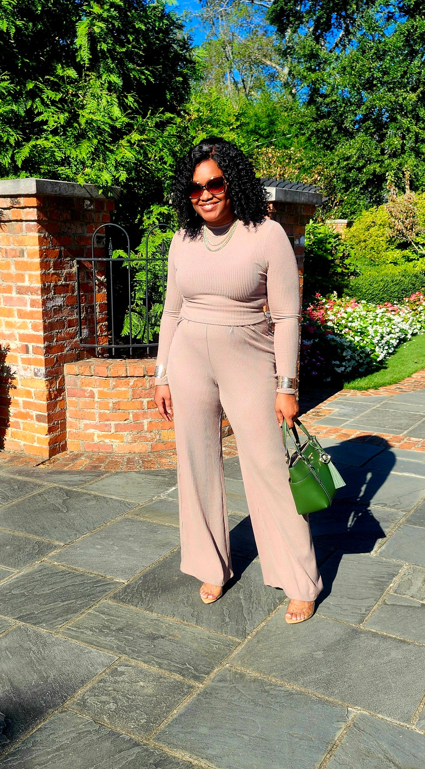Woman in a beige jumpsuit standing on a stone path with greenery in the background