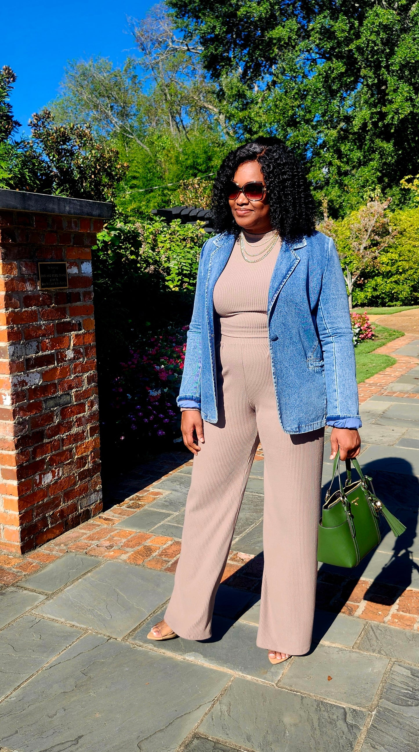 Woman in a blue blazer and beige pants standing outdoors with trees and a brick wall in the background.